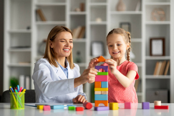 Uczenie się przez zabawę. Cute Little Girl and Child Development Specialist gra drewniane cegły podczas sesji terapeutycznej w biurze, Happy Female Kid poprawy umiejętności i cieszyć się grą, zbliżenie - Zdjęcie, obraz
