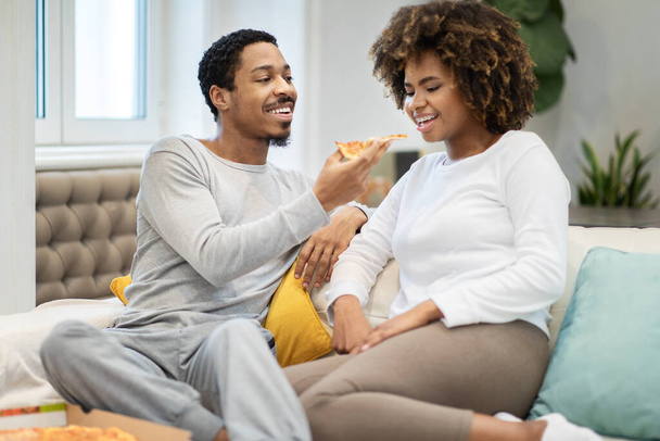 Αγαπώντας όμορφο νεαρό μαύρο άντρα που ταΐζει την κοπέλα του, ευτυχισμένο αφροαμερικάνικο ζευγάρι που κάθεται στον καναπέ στο σπίτι, τρώγοντας νόστιμη πίτσα μαζί, αντιγράφοντας χώρο - Φωτογραφία, εικόνα