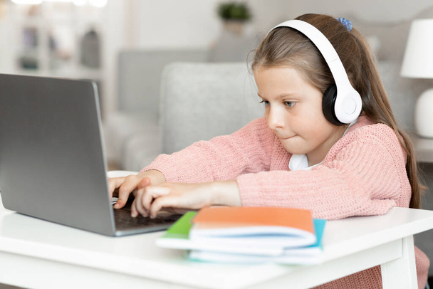 Busy concentrated caucasian small girl in headphone types, do homework, watches video lesson on computer in room interior. Childhood, learn, knowledge and education, online game at home with device - Photo, Image