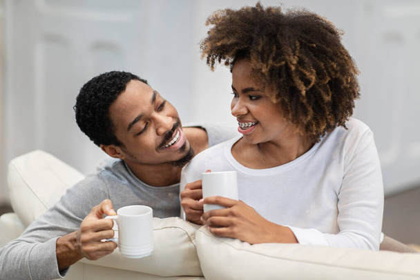 Closeup of cheerful happy young black couple drinking coffee at home. Joyful millennial african american man and woman wearing pajamas sitting on couch in living room, enjoying tea, chatting - Photo, Image