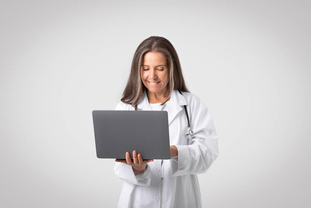 明るい灰色の背景にラップトップを使用して聴診器で白いコートで幸せな自信を持ってヨーロッパのシニア女性医師。専門的な仕事、遠隔医療、試験、広告やオファー - 写真・画像