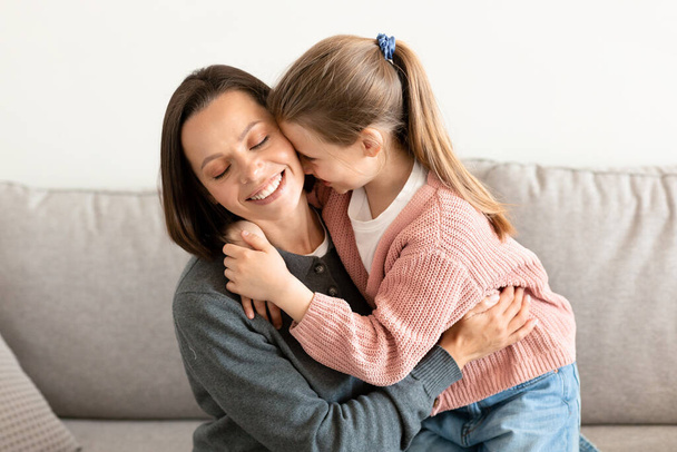 Cheerful caucasian middle aged mother hugs little daughter, enjoy tender moment in weekend in living room interior. Positive lifestyle, love, relationship family and communication at home - Photo, Image