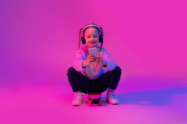 Adorable cute preteen little girl sitting on skateboard, using wireless headphones, holding cell phone, chatting with friends, scrolling on social media, luminous background. Childhood concept - Фото, изображение