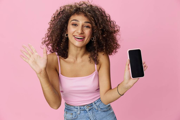 Woman blogger holding phone video call takes selfies, with curly hair in pink smile t-shirt and jeans poses on pink background, copy space, technology and social media, online. High quality photo - Photo, Image