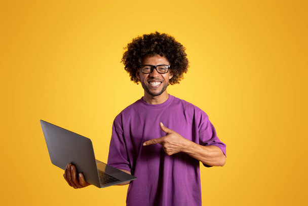 Lächelnder älterer, schwarzgelockter Mann in lila T-Shirt und Brille zeigt mit dem Finger auf Laptop auf gelbem Hintergrund, Studio. Empfehlungs- und Präsentationscomputer für Arbeit und Studium, Anzeige und Angebot - Foto, Bild