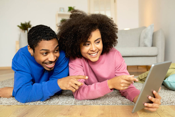 Konfor ve Teknoloji. Heyecanlı Afrikalı Amerikalı Çift Tablet Bilgisayar kullanarak Evdeki Yerde Uzanırken Dijital Keşfe ve Eğlenceye Başlıyor. Hafta sonu Web Sörf Konsepti - Fotoğraf, Görsel
