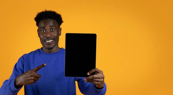 Vrolijk lachende knappe jonge zwarte man in blauwe trui wijzend naar moderne gadget digitale tablet met blanco scherm in zijn hand, mockup, gele studio achtergrond, lege ruimte. Leuke mobiele app - Foto, afbeelding