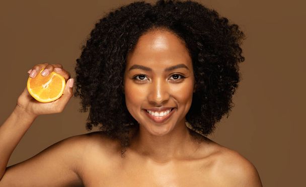 Healthy eating dieting. Vitamin detox. Cheerful half-naked young black woman holding orange for rejuvenation, skin and hair hydration, beauty treatment isolated on brown background, closeup - Photo, image