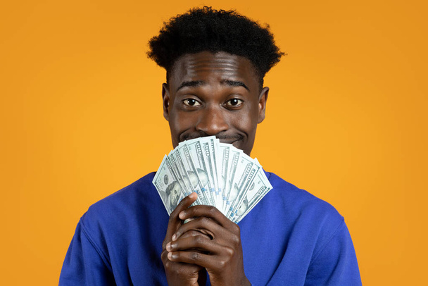 Piyango şansı. Mutlu Afrikalı Amerikalı genç adam piyangoyu kazandı. Elinde nakit para dolu banknotlar ve gülücükler var. Sarı stüdyo arka planında izole edilmiş, yakın plan. - Fotoğraf, Görsel