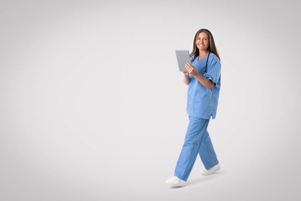 Friendly senior woman in medical uniform holding digital tablet while walking on grey background, full length, free space. Doc using medical mobile app. Telemedicine concept - Foto, Imagem