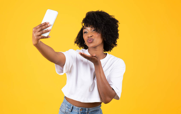 Happy young african american curly lady make blowing kiss and selfie on smartphone, have fun, isolated on yellow studio background. Social networks, beauty care, blog and photo app - Fotografie, Obrázek