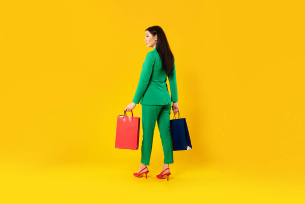 Alışveriş ve satış konsepti. Sarı arka planda izole bir şekilde duran, kağıt torbaları taşıyan bir bayanın arka görüntüsü, stüdyo çekimi, tam boy, serbest alan - Fotoğraf, Görsel