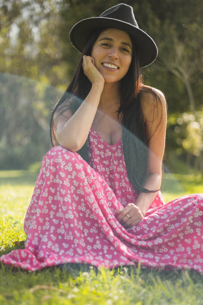 cheerful and natural young woman sitting on the grass in a summer day, she wears a beautiful long vetdio with print, beauty and youth fashion with hat, nature - Fotografie, Obrázek
