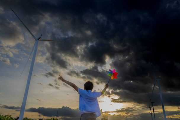 Progressive young asian boy playing with wind pinwheel toy in the wind turbine farm, green field over the hill. Green energy from renewable electric wind generator. Windmill in the countryside concept - Zdjęcie, obraz