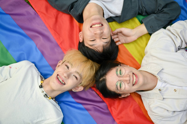 Top view of smiling and joyful young diverse Asian friends laying on a LGBT rainbow flag together. LGBTQ+. pride month, freedom, human rights - Photo, image