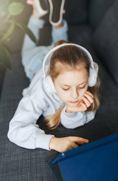 Pieni tyttö makaa sohvalla elektroninen tabletti kuuntelee musiikkia valkoiset kuulokkeet. Langattomien laitteiden käyttö koulutuksessa. - Valokuva, kuva