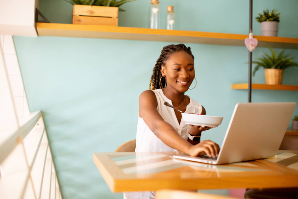 Güzel siyah kadın kafede dizüstü bilgisayarla çalışırken sağlıklı bir kahvaltı yapıyor. - Fotoğraf, Görsel