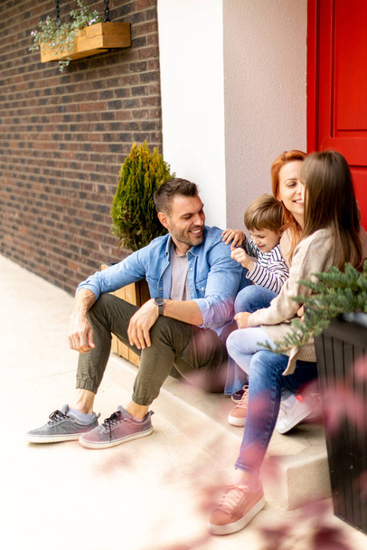 Family with a mother, father, son and daughter sitting outside on steps of a front porch of a brick house - 写真・画像
