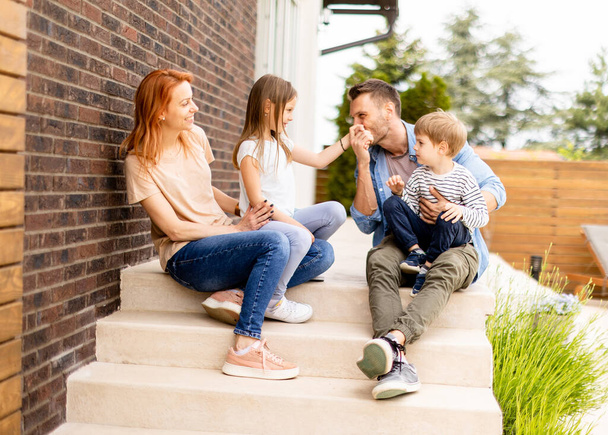 Сім'я з матір'ю, батьком, сином і дочкою, що сидить надворі на сходах переднього ганку цегляного будинку
 - Фото, зображення