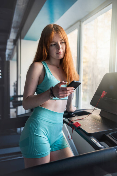 Jonge roodharige vrouw staat in de buurt van loopband trainers met behulp van mobiele cel in de sportschool. Workout, levensstijl - Foto, afbeelding