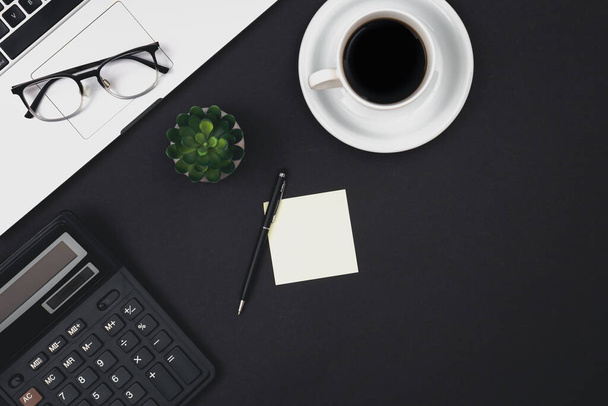 Laptop, brýle, šálek kávy a kalkulačka na černém pozadí, pohled shora, minimalistické zázemí, pracoviště. - Fotografie, Obrázek