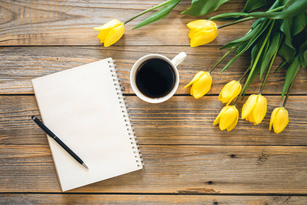 A blank notebook, a cup of black coffee and yellow tulips on a wooden background, top view. - Фото, изображение