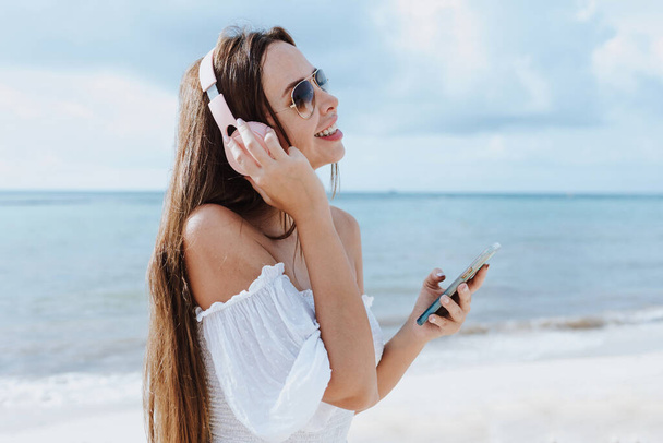 携帯電話を使用してリラックスしたラテン女性とメキシコのカリブ海ビーチで音楽を聴くヘッドフォンを身に着けているラテンアメリカ、ヒスパニック女性  - 写真・画像