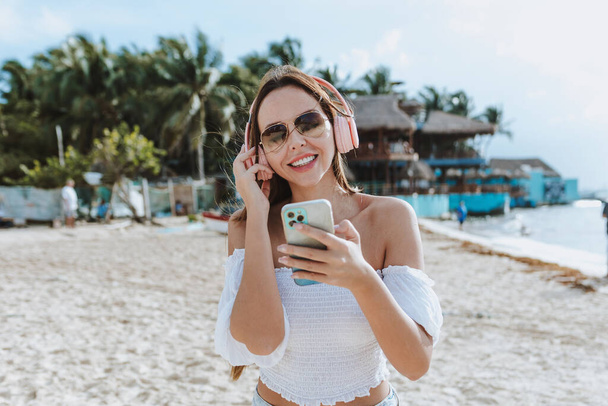 Relaxed Latin woman using mobile phone and wearing headphones listening to music at Caribbean Beach in Mexico Latin America, hispanic female  - Foto, Imagem