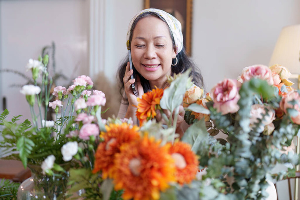 Indítás, kisvállalkozás, virágüzlet. Az idősebb virágárus virágot tesz egy vázára, és telefonon beszél a vevővel.. - Fotó, kép