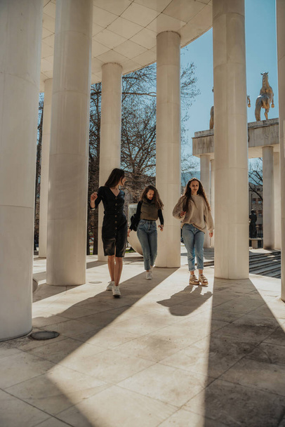 Girls hanging out in the city. Brunette girl in black overcoat walking forward while looking back at her friends and talking to them - Photo, image