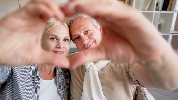 Love message. Family couple. Lovely moments. Happy middle-aged man and woman showing heart gesture combining hands together in light home interior. - Φωτογραφία, εικόνα