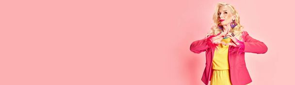 Portret van een mooie oudere blonde vrouw met heldere make-up, in kleurrijke levendige kleren poseren tegen roze studio achtergrond. Concept van schoonheid, mode, menselijke emoties, levensstijl. Een spandoek. Kopieerruimte - Foto, afbeelding