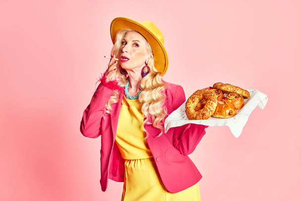 Portrait of stylish, beautiful, senior woman in colorful clothes smoking cigarette and holding fresh sweet buns against pink studio background. Concept of beauty, fashion, human emotions, lifestyle - Foto, Bild