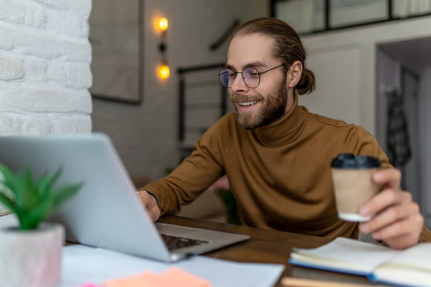 Young smiling bearded man freelancer in glasses working at home or in cafe with laptop drinking takeaway coffee. - Fotoğraf, Görsel