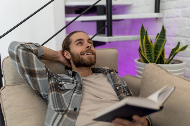 Handsome smiling happy young man reading book and dreaming while sitting on sofa in his living room. - Φωτογραφία, εικόνα