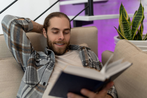 Cheerful relaxed man reading book while sitting on cough at home, enjoying interesting novel. - Foto, immagini