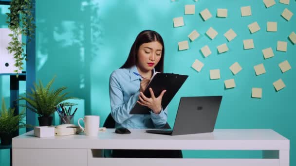 Γραμματέας γραφείου συμπληρώνοντας λίστα ελέγχου στο πρόχειρο με βάση τις πληροφορίες που λαμβάνονται από το διαχειριστή κατά τη διάρκεια σε απευθείας σύνδεση videocall. Υπάλληλος γυναίκα σε πολύχρωμο γραφείο πάνω από το μπλε φόντο στούντιο - Πλάνα, βίντεο