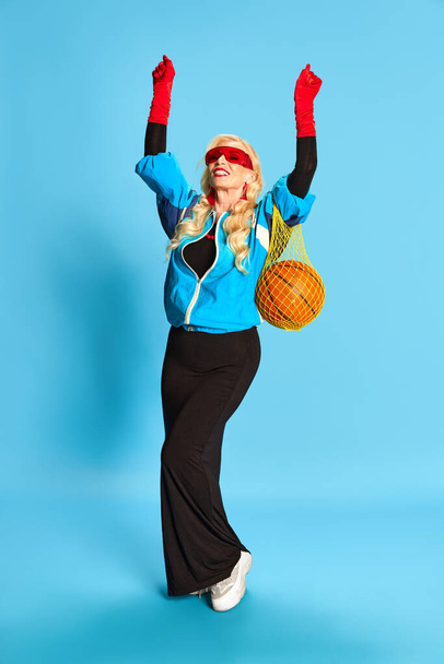 Full-length portrait of smiling senior woman in black dress and sport jacket posing with basketball ball against blue studio background. Concept of beauty, fashion, human emotions, lifestyle. Ad - Фото, изображение