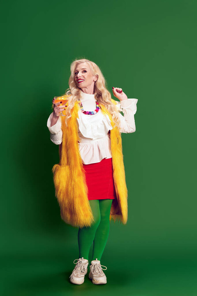 Portrait of beautiful senior woman in stylish clothes, yellow fur coat drinking cocktail against green studio background. Party time. Concept of beauty, fashion, human emotions, lifestyle. Ad - Φωτογραφία, εικόνα