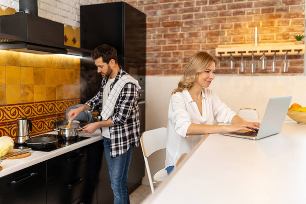 Happy healthy relationship. Woman working on laptop in kitchenn while boyfriend preparing meal. - Foto, Bild