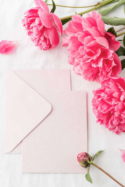 Peony flowers and blank light pink paper post card with open envelope on white linen table cloth background. Invitation floral card or letter in vintage style. Mockup birthday greeting card. - 写真・画像