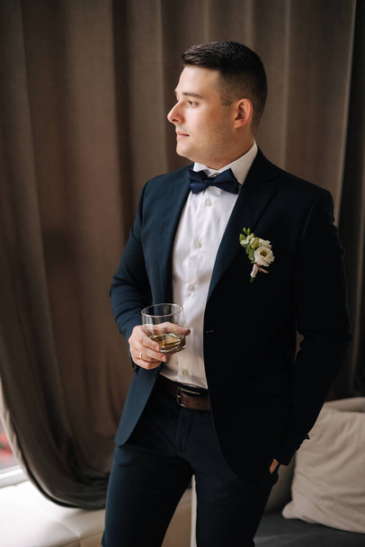 Young groom stand near big window and hold glass of whiskey at home. Morning of groom at wedding day. High quality photo - Fotoğraf, Görsel