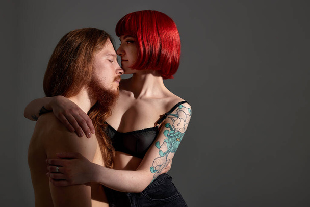 A young attractive woman with bright ruby hair and a tattoo in black lingerie embraces a man with long hair and a beard. Half-naked lovers in each others arms. Close-up studio shot, dark background - Photo, Image