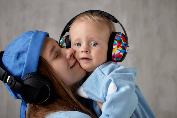 Joyeux jeune mère tient et étreint son bébé souriant. Le bébé et la mère portent des vêtements bleus similaires et des écouteurs DJ drôles sur la tête. Plan studio, fond gris. - Photo, image