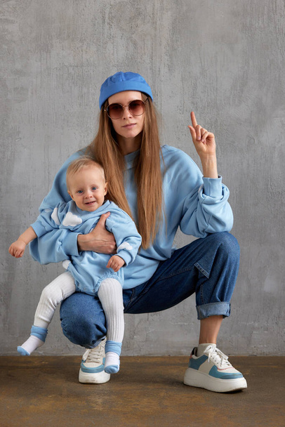Een jonge stijlvolle mama in spijkerbroek, een blauwe trui, een grappige blauwe pet en een zonnebril zit op haar broek en knuffelt haar lachende baby. Baby en mama dragen dezelfde kleur kleren. Studio shot, grijze achtergrond - Foto, afbeelding