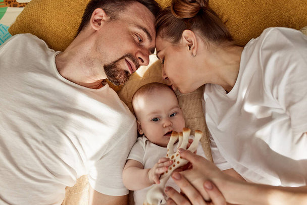かわいい喜びの赤ちゃんと彼の両親はおもちゃで明るい居心地の良い部屋に床に横たわっています。お母さんとお父さんは抱き合って赤ちゃんにキス。幸せな親と子供時代 - 写真・画像