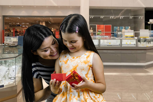 A menina abre uma caixa de presente vermelha da mãe. Conceito de celebração de aniversário. Mulher e sua filha no shopping. - Foto, Imagem
