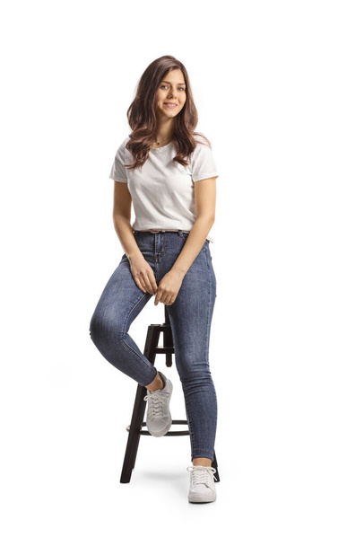 Beautiful young woman in jeans and white t-shirt sitting on a high chair isolated on white backgroun - Foto, Bild