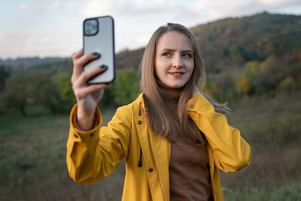 Mujer joven en chaqueta de parque amarillo disfruta de la belleza de la naturaleza otoñal.Retrato de chica sonriente tomando selfie en el teléfono inteligente en el fondo del paisaje de montaña - Foto, Imagen