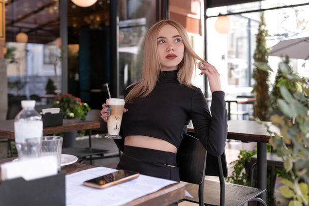 Brooding blonde woman sits in coffee shop with cup of cappuccino coffee latte. Attractive girl alone in cafe. - Fotoğraf, Görsel
